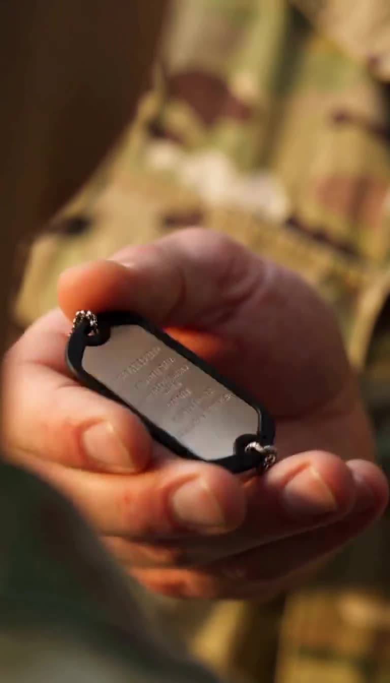 Extreme close-up of Staff Sergeant Marcus's hand flipping the dog tag to reveal the engraving