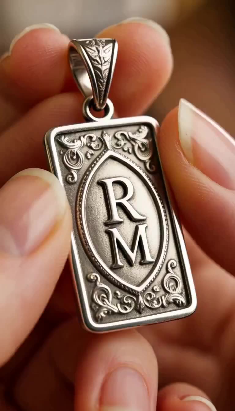 Close-up of fingers holding a silver rectangular pendant engraved with the letters R and M