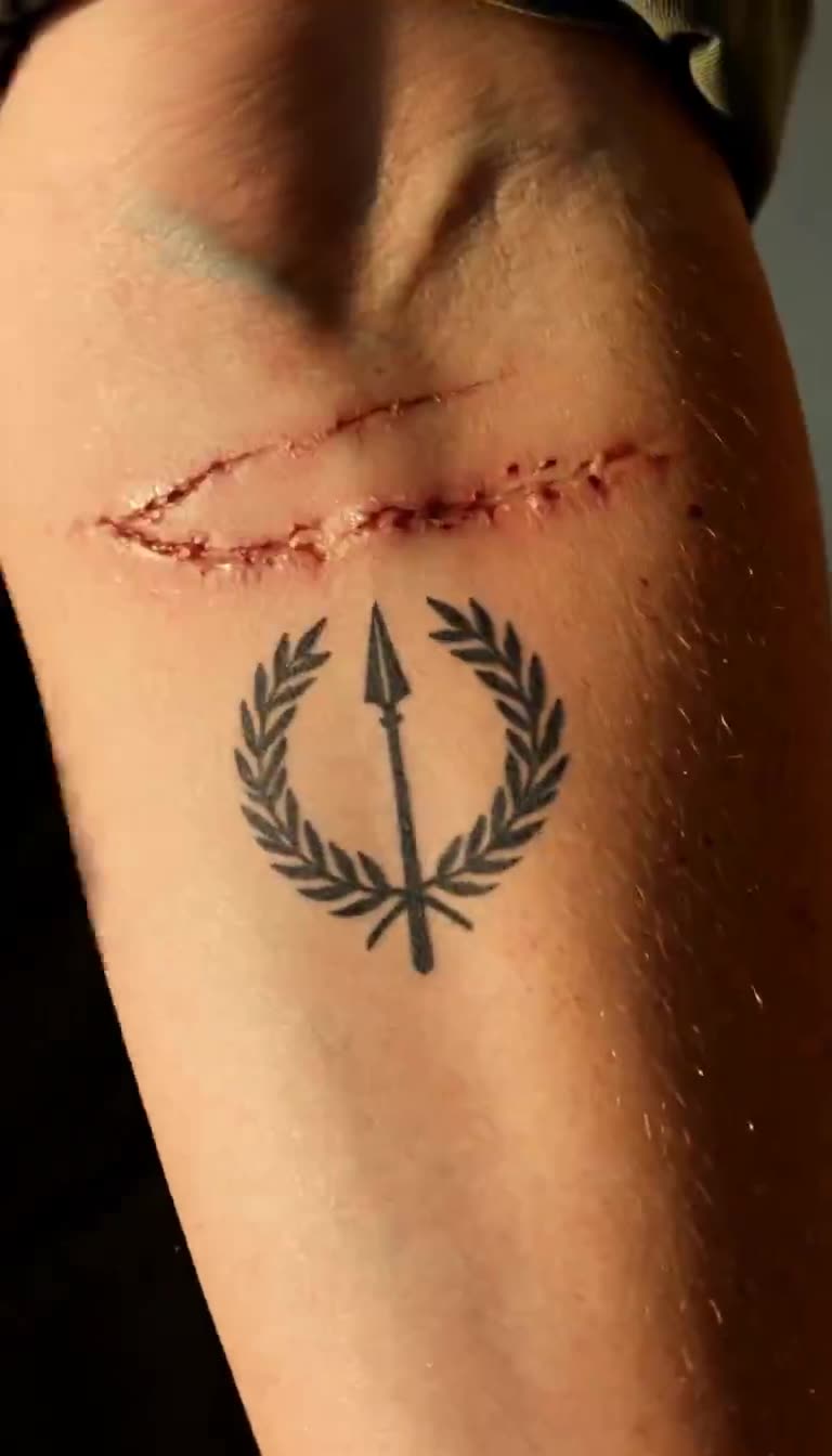 Extreme close-up of healed scars and a spear with laurel wreath tattoo on forearm