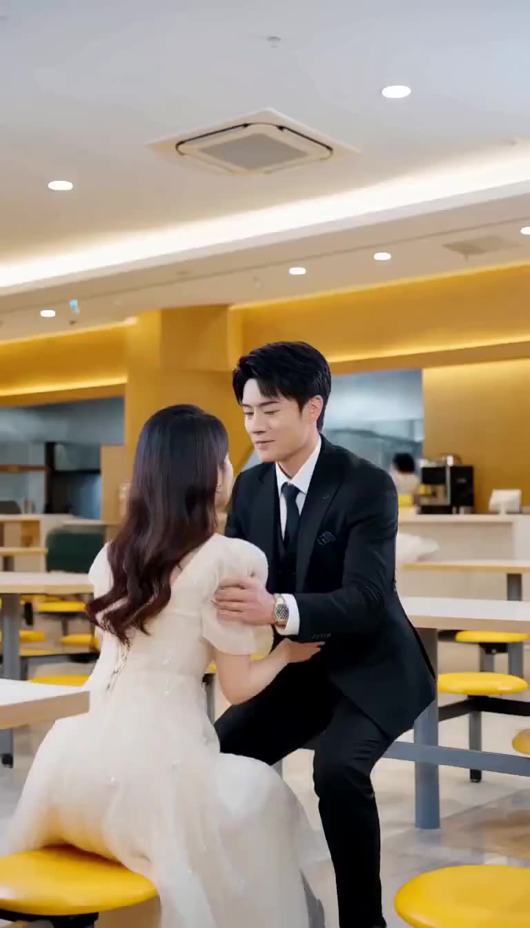 A man in a dark suit holding the shoulders of a woman in a white dress seated in a bright canteen with yellow stools